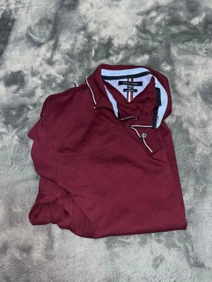 Tommy Hilfiger Slim Fit Polo - en riktigt fräsch pikétröja från Tommy Hilfiger i en snygg vinröd färg. Den har snygga kontrastränder vid kragen och knapparna som ger det där lilla extra. Sitter riktigt snyggt och följsamt på kroppen
