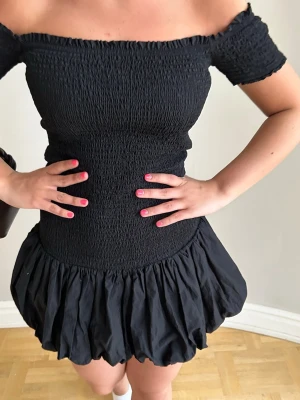 Svart offshoulder kort klänning💖💖🌸 - Trendig svart kort klänning med offshoulder och smockad överdel. Kjolen är volangig och ger en lekfull look. Perfekt för dig som gillar en feminin och stilren stil. Klänningen har en figurnära passform upptill och är tillverkad i ett lätt material.