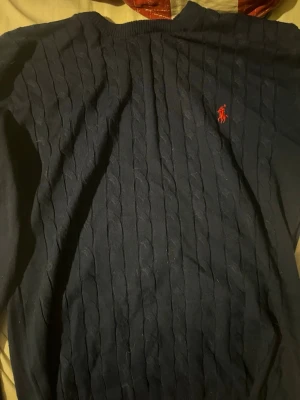Mörkblå kabelstickad tröja Polo Ralph Lauren - Klassisk mörkblå kabelstickad tröja från Polo Ralph Lauren med röd broderad logga på bröstet. Tröjan har rund halsringning och långa ärmar. Tillverkad i mjukt material som passar perfekt till lager på lager-stil.
