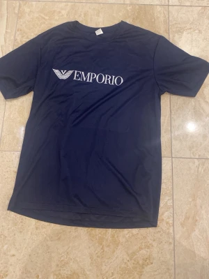 Mörkblå Emporio t-shirt med tryck - Snygg mörkblå t-shirt från Emporio med vitt logotryck på bröstet. Klassisk passform med rund hals och korta ärmar. Perfekt för en stilren och enkel look.