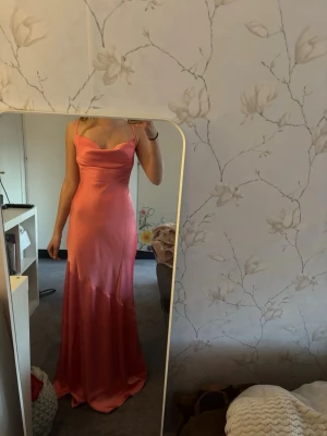 Lång rosa satinklänning med smala axelband - Rosa långklänning i satin med smala axelband och lätt draperad urringning. Klänningen har figurnära passform och faller snyggt längs kroppen. Perfekt för speciella tillfällen såsom en bal