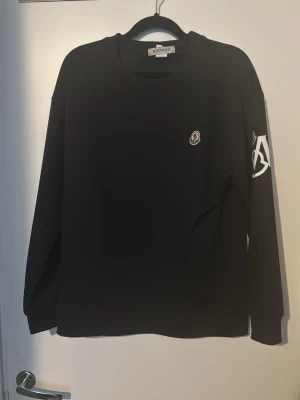 Moncler tröja - Svart sweatshirt från Moncler med rund halsringning och lång ärm. Liten Moncler-logga på bröstet och vitt tryck på ena ärmen. Perfekt för en stilren och trendig look. Använd 2 gånger inga defekter, storlek L men passar M