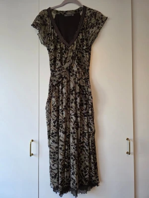 Lång blommig klänning med spetsdetaljer - Snygg klänning med svart blommönster på beige-grön botten. Djup V-ringning med dekorativ spetskant och korta ärmar. Klänningen har ett lätt, flytande tyg och markerad midja. Töjbar.