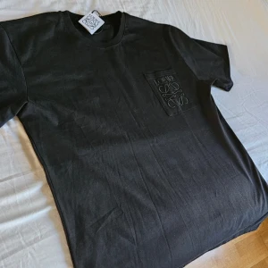 Svart t-shirt från Loewe med ficka - Säljer en svart t-shirt från Loewe med en bröstficka där Loewe-loggan är broderad ton-i-ton. Klassisk rund halsringning och korta ärmar. Perfekt för dig som gillar stilrena och exklusiva plagg. Premium tyg. Känns lyxigt! Pris går att diskutera!