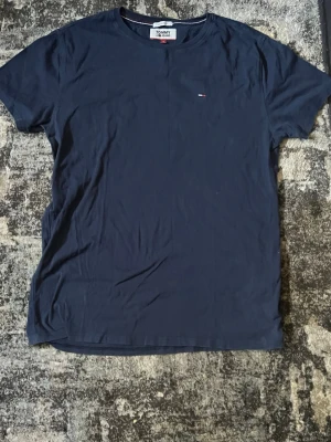 Mörkblå t-shirt från Tommy Jeans - En stilren mörkblå t-shirt från Tommy Jeans med rund halsringning och liten broderad logga på bröstet. Tillverkad i mjuk bomull och har en klassisk passform som funkar till det mesta. Perfekt för dig som gillar enkel och snygg stil.