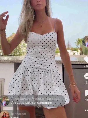 Vit prickig kort klänning - Söt vit kort klänning med svarta prickar och tunna axelband. Klänningen har en figurnära överdel och en utställd kjol som ger en lekfull look. Perfekt för varma dagar och sommarhäng.