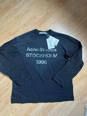 Svart långärmad t-shirt Acne Studios - Svart långärmad t-shirt från Acne Studios med tryck 'Acne Studios STOCKHOLM 1996' på bröstet. Klassisk passform och rund halsringning. Tillverkad i mjuk bomull, perfekt för en stilren och avslappnad look.
