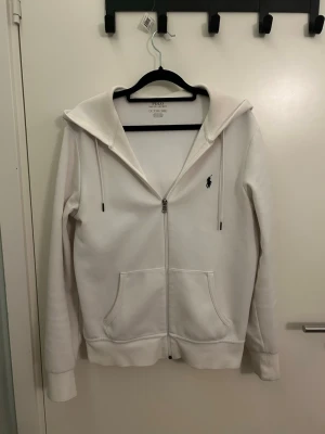 Vit hoodie från Ralph Lauren - Snygg vit hoodie från Ralph Lauren med dragkedja framtill och klassisk broderad logga på bröstet. Hoodien har huva med dragsko och två stora fickor framtill. Perfekt för en avslappnad och stilren look. Storlek XS men passar S
