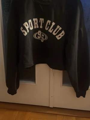Svart cropped hoodie Gina Tricot - Mörk grå cropped hoodie från Gina Tricot med trycket 'SPORT CLUB' i vitt framtill. Tröjan har långa ärmar och ribbade muddar. Materialet är mjukt och bekvämt. Knappt använd. Säljer då jag inte använder den längre.