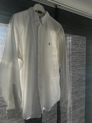 Vit skjorta från Ralph Lauren - Vit klassisk skjorta från Ralph Lauren med broderad logga på bröstet. Skjortan har lång ärm, knappar framtill och button-down krage. Materialet är bomull och passformen är classic fit. Perfekt för dig som gillar stilrena plagg.