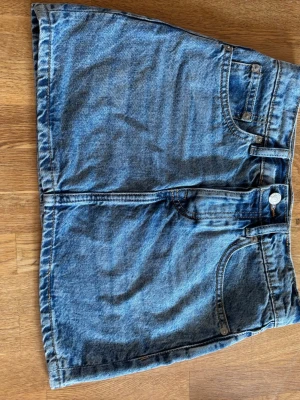 Blå jeanskjol i klassisk modell - Snygg blå jeanskjol från 157 med klassisk femficksdesign, bälteshällor och knappstängning framtill. Kjolen är kort och har en rak passform, perfekt till vardagsstilen. Tillverkad i denim för en tidlös look.