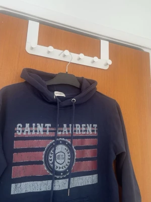 Mörkblå Saint Laurent hoodie - Snygg mörkblå hoodie från Saint Laurent med tryck i rött, vitt och blått framtill. Klassisk huva med dragsko och ribbade muddar. Perfekt för en avslappnad och trendig stil. Materialet känns mjukt och bekvämt mot huden.