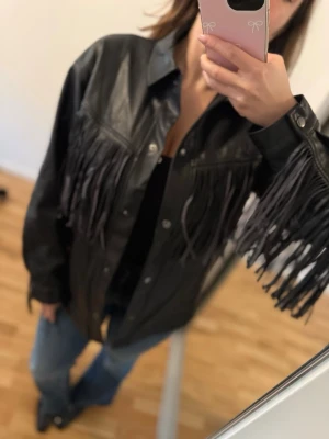 Svart skinnjacka med fransar - Cool svart skinnjacka med långa fransar över bröstet och längs ärmarna. Jackan har klassisk krage och knäppning framtill. Perfekt för dig som vill sticka ut med en trendig westernstil.