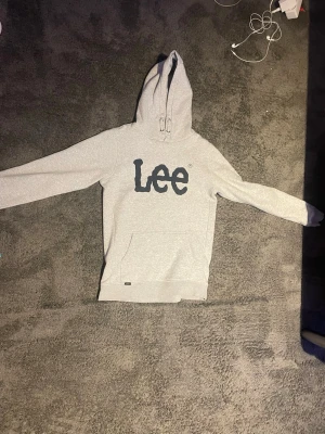 Grå hoodie från Lee med tryck - Snygg ljusgrå hoodie från Lee med stort svart tryck på bröstet. Klassisk modell med huva och känguruficka framtill. Perfekt för en avslappnad streetstyle-look. Mjuk bomullsblandning och ribbade muddar vid ärmslut och nederkant. Igentliten storlek 14-15 år men fanns inte att lägga in
