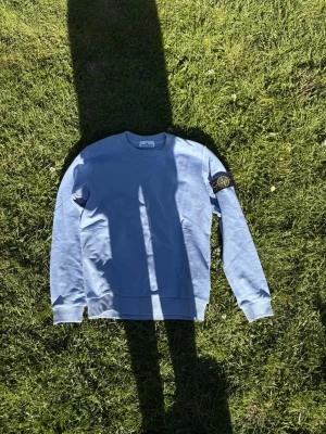 Ljusblå stickad tröja Stone Island - Ljusblå stickad tröja från Stone Island med klassisk ribbad krage och ärmslut. Tröjan har den ikoniska svarta patchen med gult broderi på ärmen. Perfekt för dig som gillar streetwear och snygga detaljer.