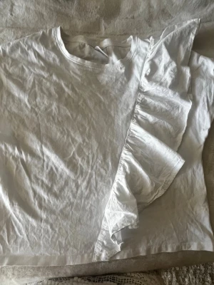 Vit t-shirt med volangdetalj - Säljer en vit t-shirt med rund halsringning och en snygg volangdetalj som går diagonalt över framsidan. T-shirten har korta ärmar och är gjord i ett mjukt bomullstyg. Perfekt för dig som gillar enkla plagg med en twist. Den ser skrynklig ut eftersom att jag inte har strikt den, jättesnygg på. 