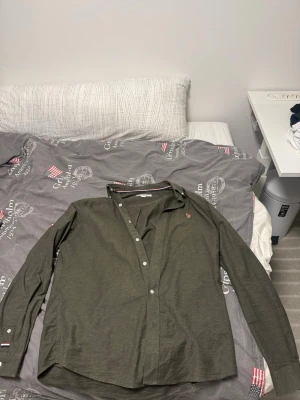 Olivgrön skjorta från Tommy Hilfiger - Snygg olivgrön skjorta från Tommy Hilfiger med klassisk krage och långa ärmar. Skjortan har knappar framtill och små logodetaljer på ärmsluten. Perfekt för en stilren look. Den har knappt använts. Äkta polo.
