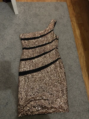Glittrig one shoulder klänning - Snygg glittrig kortklänning med paljetter i guld och svarta diagonala ränder. Klänningen har en oneshoulder-design och är figurnära, perfekt för dig som vill sticka ut. Materialet är syntet med paljett-detaljer.