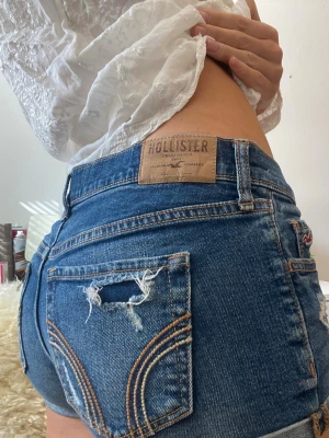 Blå denimshorts från Hollister - Snygga blå denimshorts från Hollister med låg midja och slitna detaljer på bakfickan. Klassisk femficksmodell med uppvikta benslut och tydliga kontrastsömmar. Perfekta för varma dagar och enkel att matcha med allt.