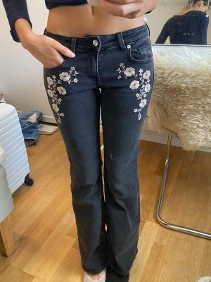 Svarta bootcut jeans med blommor - Snygga svarta bootcut jeans med vita och ljusrosa broderade blommor framtill vid fickorna. Jeansen har klassisk femficksmodell och stängs med knapp och dragkedja. Perfekta för dig som vill ha en unik och trendig look.