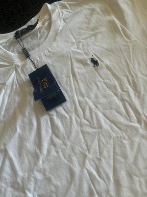 Vit t-shirt från Polo Ralph Lauren - Klassisk vit t-shirt från Polo Ralph Lauren med rund halsringning och ikoniska broderade loggan på bröstet. Tillverkad i mjuk bomull som känns skön mot huden. Perfekt basplagg med stilren design och diskreta detaljer.