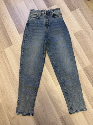 Blå mom jeans med hög midja - Säljer ett par klassiska blå mom jeans med hög midja och raka ben. Jeansen har fem fickor, dragkedja och knapp framtill. Perfekta för en avslappnad och trendig stil. Materialet är 100% bomull för en autentisk denimkänsla.