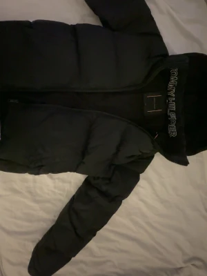 Svart pufferjacka från Tommy Hilfiger - Snygg svart pufferjacka från Tommy Hilfiger med huva och dragkedja. Jackan har broderad logga på insidan av kragen och är vadderad för extra värme. Perfekt för kalla dagar och har en klassisk, stilren look. storlek 168cm