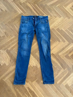 Replay anbass blå jeans - Säljer ett par blå slim fit jeans från Replay med klassisk femficksdesign. Jeansen har dragkedja och knapp i midjan samt detaljerade bakfickor. Perfekta för en avslappnad och trendig look. Litet hål syns på bild 3 men bild 5 visar att det är lagat inifrån. Storlek 32/34