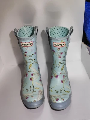 Ljusblå blommiga Hunter gummistövlar - Säljer ett par ljusblå gummistövlar från Hunter med blommigt mönster i rosa, gult och grönt. Stövlarna har handtag upptill och prickigt foder på insidan. Perfekta för regniga dagar och festivaler! Storlek 35/36