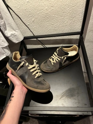 Bruna sneakers från Maison Margiela - Säljer ett par bruna sneakers från Maison Margiela med beige sula och vita snören. Skorna har en klassisk low-top siluett med ovandel i mocka och läder, samt diskret logga på plösen. Perfekta för dig som gillar stilrena och tidlösa sneakers.