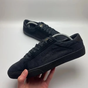 Saint Laurent SL/06 Triple Black - Snygga helsvarta sneakers från Saint Laurent med diskret logga på sidan och gulddetaljer på plösen. Skorna har en klassisk låg siluett, ovandel i mocka och textil samt matchande svarta skosnören. Perfekta för en clean och stilren look.