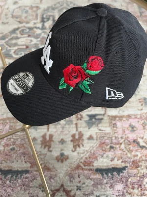 Svart LA-keps med rosbrodyr - Snygg svart keps från New Era, modell 9FORTY, med LA-logga i vitt framtill och två röda rosor broderade på sidan. Justerbar passform och klassisk böjd skärm. Materialet är bomullstyg och kepsen har en sportig streetwear-vibe.