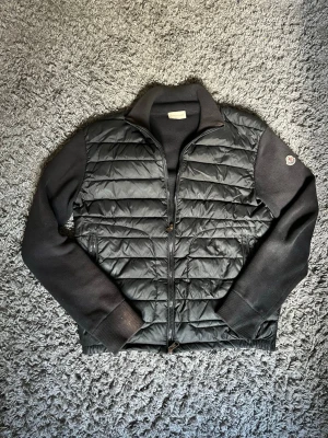 Moncler cardigan - Säljer en svart cardigan från Moncler med quiltad framsida och stickade ärmar samt rygg. Jackan har hög krage, dragkedja och Moncler-logga på ena ärmen. Perfekt för en lite kyligare sommarkväll och riktigt snygg att bära till jeans.