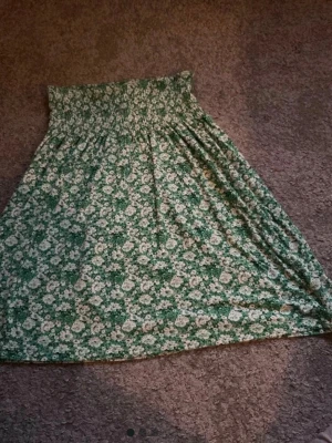 Grön blommig långkjol från Indiska - Grön långkjol från Indiska med vitt blommönster och smock i midjan. Kjolen har en luftig och flowy passform, perfekt för varmare dagar. Tillverkad i mjukt och lätt material som känns skönt mot huden.