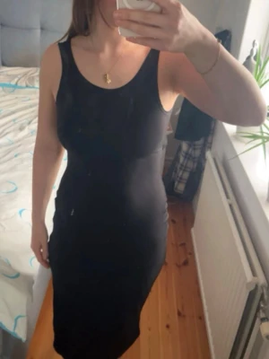 Svart ärmlös klänning med öppen rygg - Snygg svart klänning med tight passform och ärmlös design. Klänningen har en cool öppen rygg med korsade band och en rundad halsringning framtill. Perfekt för dig som gillar stilrena och trendiga plagg.