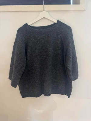 Mörkgrå stickad  tröja - Mysig mörkgrå stickad tröja med avslappnad och rund halsringning. Tröjan har trekvartsärmar och är tillverkad i ett mjukt, fluffigt material som känns skönt mot huden. Perfekt för lager-på-lager och chill dagar.