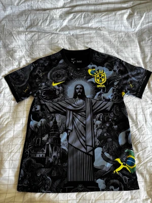 Svart Brasilien Nike Dri-Fit tröja - Snygg svart Brasilien-tröja från Nike med Dri-Fit-material. Tröjan har ett stort tryck av Kristusstatyn och detaljer i gult, inklusive Brasil-logga och Nike-swoosh. Unik design med konstnärliga mönster och korta ärmar. Perfekt för fotboll eller streetwear.