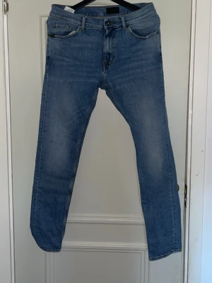 Blå slim fit jeans från Tiger of Sweden - Snygga blå slim fit jeans från Tiger of Sweden med klassisk femficksdesign och läderpatch bak i midjan. Jeansen har en något tvättad look och raka ben. Perfekta för en stilren och avslappnad outfit.