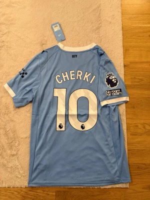 Manchester City matchtröja Cherki #10 - Ljusblå Manchester City matchtröja från Puma med Cherki och nummer 10 på ryggen. Tröjan har vita och mörkblå detaljer, klubbmärke, Premier League-logga och 'No room for racism'-patch på ärmen. Materialet är lätt och andas, perfekt för fotboll.