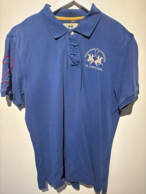 Blå pikétröja från La Martina - Snygg blå pikétröja från La Martina med broderad logga och polospelare på bröstet. Klassisk krage, korta ärmar och knappslå framtill. Röd detalj på ena ärmen. Perfekt för en sportig och stilren look.