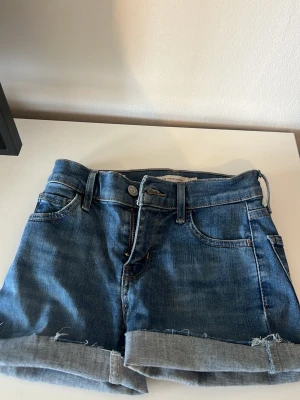 Blå jeansshorts från Levi's - Klassiska blå jeansshorts från Levi's med upprullade benslut och femficksdesign. Shortsen har normal passform, bälteshällor och synliga sömmar. Perfekta för sommaren och enkla att matcha med olika stilar.