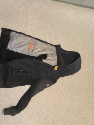 Svart jacka med huva från Parajumpers - JACKAN ÄR SOL BRÄND PÅ ARMARNA SÅ NI VET OCH DET HADE HÅL I ARMAR MEN JAG HAR SYTT IHOP HÅLEN SÅ HÅLEN SYNS INTE.... Svart jacka från Parajumpers med huva och patch på ärmen. Jackan har quiltad framsida, dragkedja och flera fickor. Insidan är grå med orange detaljer. Perfekt för kyliga dagar och har en sportig look.