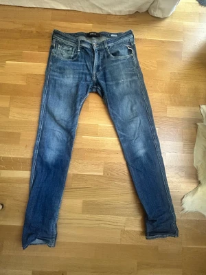 Blå slim fit jeans från Replay - Säljer ett par blå slim fit jeans från Replay med snygga slitningar och klassisk femficksdesign. Jeansen har en något tvättad look och detaljer som kontrastsömmar och läderpatch bak i midjan. Kom med frågor, priset är ej huggit i sten 