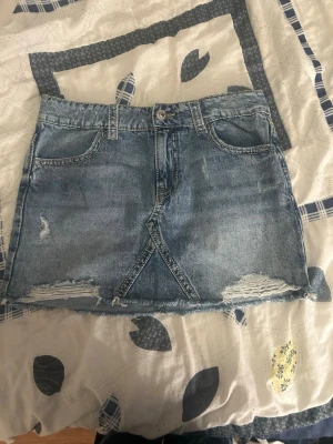 Blå jeanskjol med slitna detaljer - Snygg blå jeanskjol i kort modell med slitna detaljer och rå kant nertill. Klassisk femficksdesign och knappstängning framtill. Perfekt för dig som gillar en avslappnad och trendig stil.