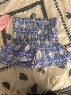 Blå mönstrad bandeau topp Gina Tricot - Supersöt bandeau topp från Gina Tricot i blått med vitt blommigt mönster. Toppen är axelbandslös och har en volang nertill som ger en extra touch. Perfekt för varma dagar och sommarvibbar!