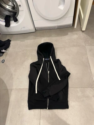 Svart hoodie från Polo Ralph Lauren - Säljer en svart hoodie med dragkedja från Polo Ralph Lauren. Hoodien har vita snören, huva och det klassiska röda Polo-logotypen broderad på bröstet. Tillverkad i mjukt bomullsmaterial och har en avslappnad passform. Perfekt för en chill och stilren look. Väldigt grich