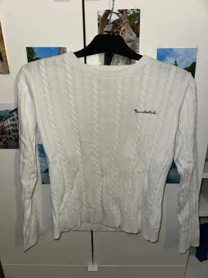 Vit kabelstickad tröja Bondelid - Snygg vit kabelstickad tröja från Bondelid med broderad logga på bröstet. Tröjan har rund halsringning, långa ärmar och ribbade muddar. Perfekt för lager-på-lager och passar till det mesta i garderoben.