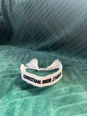 Vitt armband Christian Dior J'Adior - Snyggt vitt tygarmband från Christian Dior med texten 'J'Adior' broderad i svart. Armbandet har justerbar knytning med tofsar i ändarna och är perfekt för att ge din outfit en exklusiv touch.