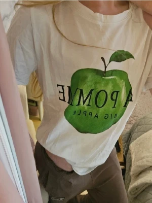 Vit t-shirt med grön äppleprint - Säljer en jätte söt vit t-shirt från H&M i storlek S med stort grönt äpple och texten 'LA POMME BIG APPLE' på framsidan. Klassisk rund hals och korta ärmar. Tillverkad i mjuk bomull. Är använd men har inga tecken på fläckar, skador eller andra slitage. Säljer den då den tyvärr inte längre kommer till användning. Nypris runt 150 kr. 💓