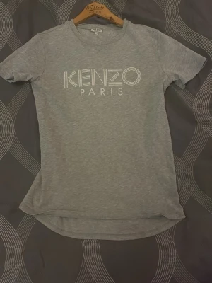 Grå Kenzo Paris t-shirt - Snygg grå t-shirt från Kenzo med vitt tryck på bröstet där det står 'KENZO PARIS'. T-shirten har rund halsringning och korta ärmar. Perfekt för en stilren och trendig look.